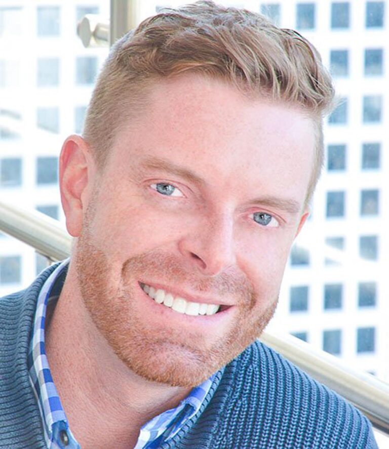 The image shows a man in a blue and white checkered shirt under a blue sweater. The background shows a window overlooking a building that has a grid-like pattern of windows.