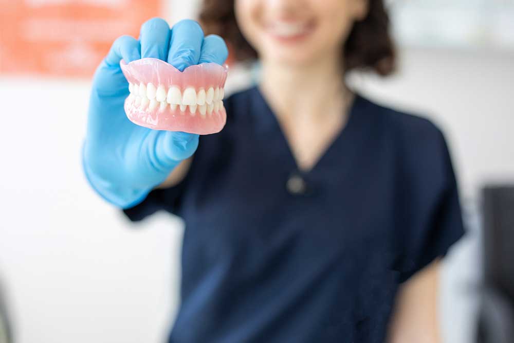 The image displays a pair of dentures held by a person in a dark blue uniform and blue gloves. The dentures, featuring pink gums and white teeth, are close to the camera and emphasize dental prosthetics, with a blurred dental office setting in the background.