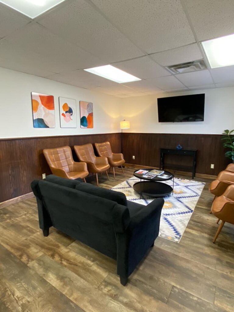 Modern waiting room with wooden floors and white and dark wood-paneled walls. Three leather chairs face a black couch with a glass coffee table on a patterned rug. Decor includes abstract art, a wall-mounted TV, potted plant, and floor lamp. The ceiling features recessed lighting and air vents.